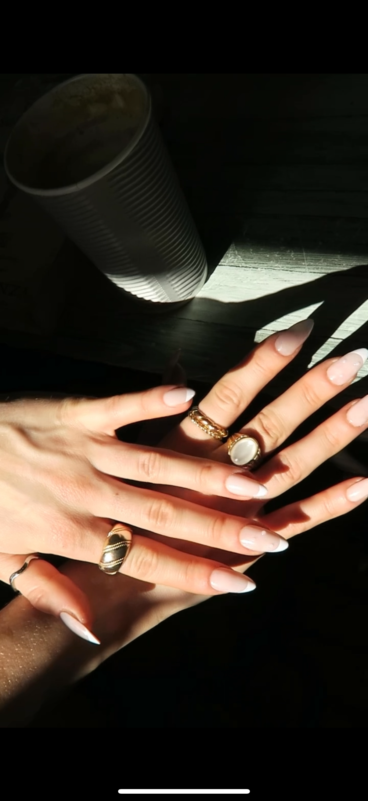 Close-up of hands with multiple rings on a dark background wearing French tip press on nails with pearl detail.