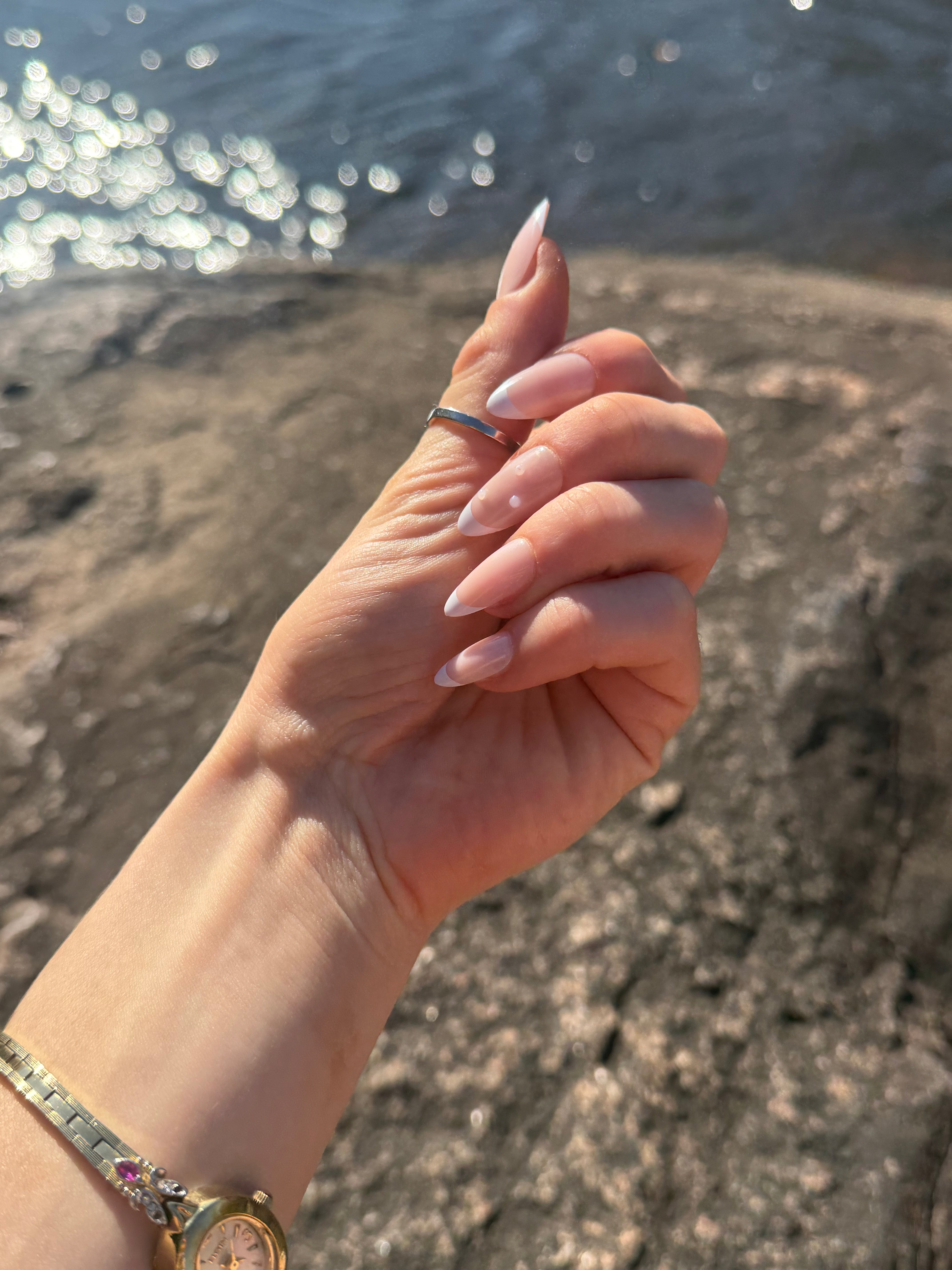 Hand with a ring on a beach wearing French tip press on nails with pearl detail.