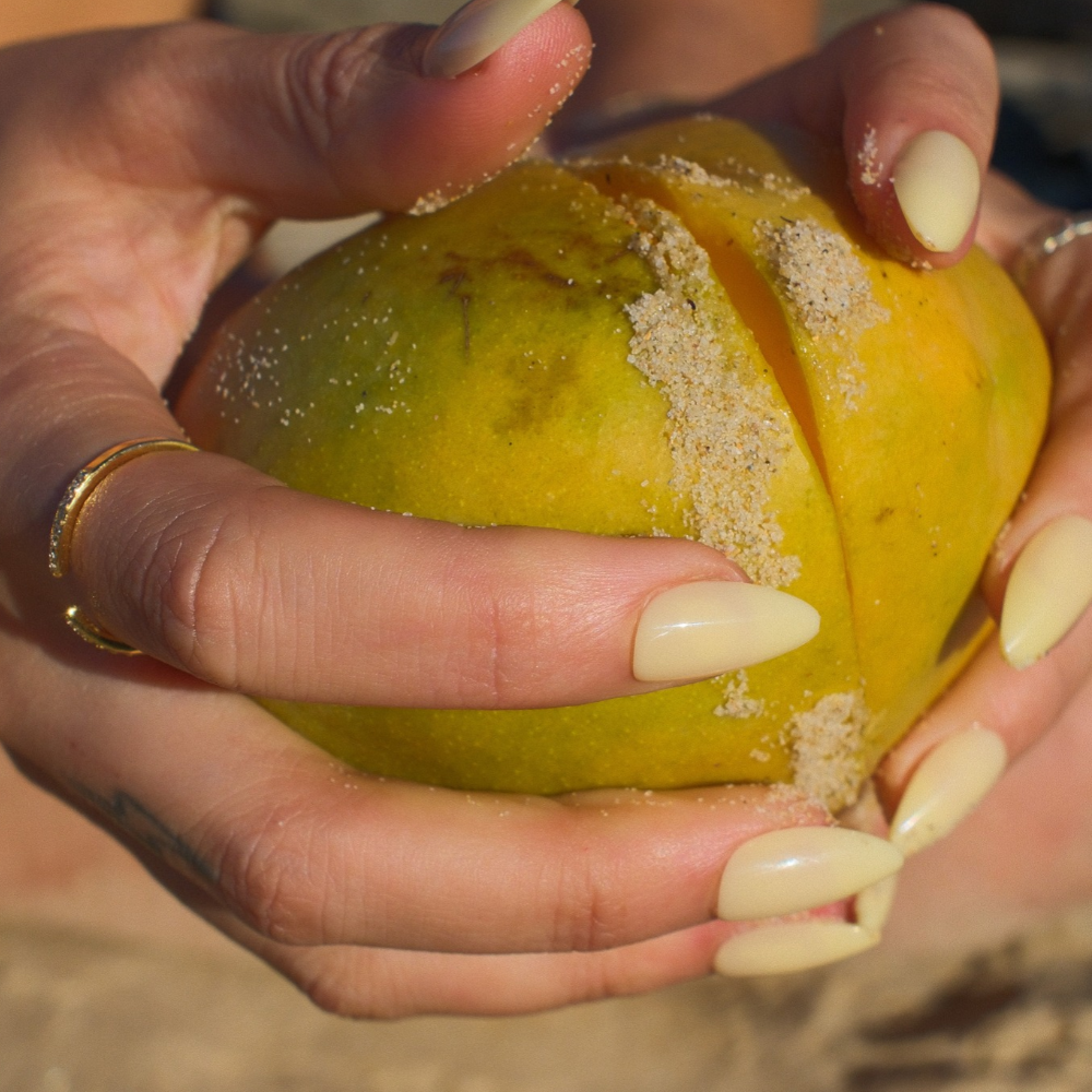 Butter Glow butter yellow press-on nails in medium almond by Sugar Tips, holding a sandy mango at the beach
