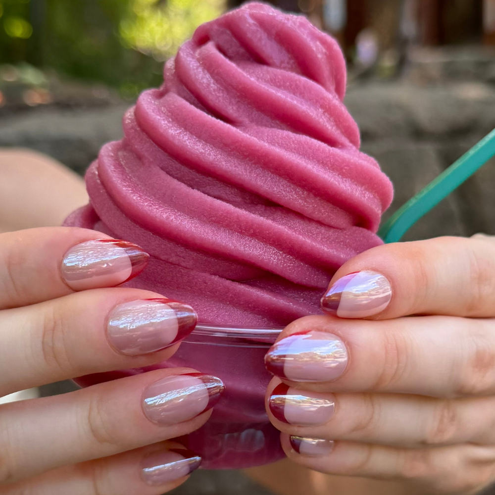 A close-up of a hand holding a pink frozen dessert or soft serve in a clear cup, with press on nails from Sugar Tips showing a sheer nude base and iridescent shimmer with red French tips.