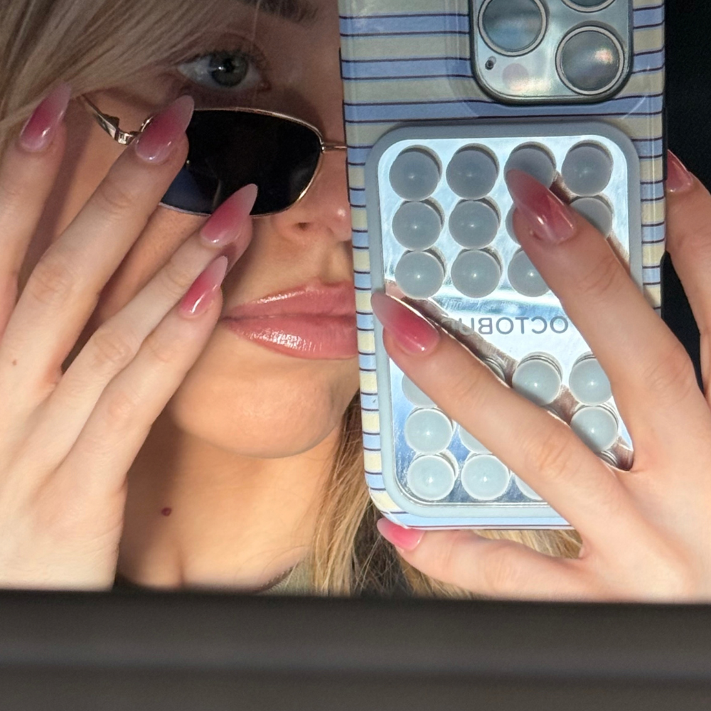 A close-up selfie showing a person holding a phone with a clear case containing round gel pads. The person is also wearing dark sunglasses and has long pink ombré press on nails from sugar tips.