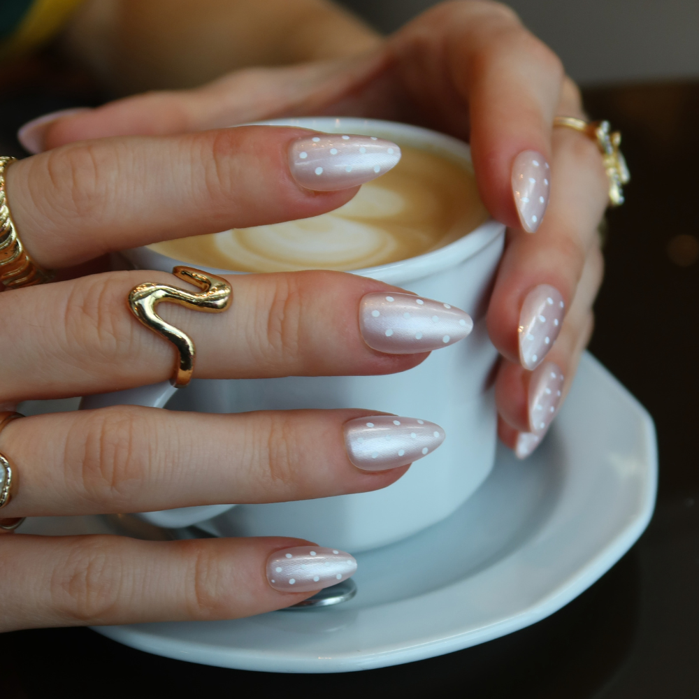 A close-up of hands holding a white coffee cup on a saucer. The nails have on Press On Nails from Sugar Tips in a long, almond-shape, and decorated with a pale pink glitter base and small white polka dots.