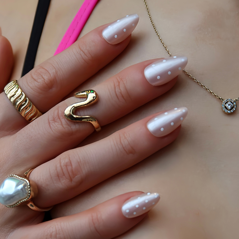 A close-up of a hand with long, almond-shaped Press On Nails decorated with small white polka dots, styled with multiple gold rings and a delicate gold necklace on a beige skin background.