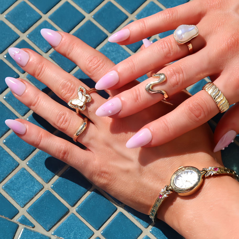 Lilac almond press-on nails by SugarTips, worn with gold rings and a watch against blue pool tiles