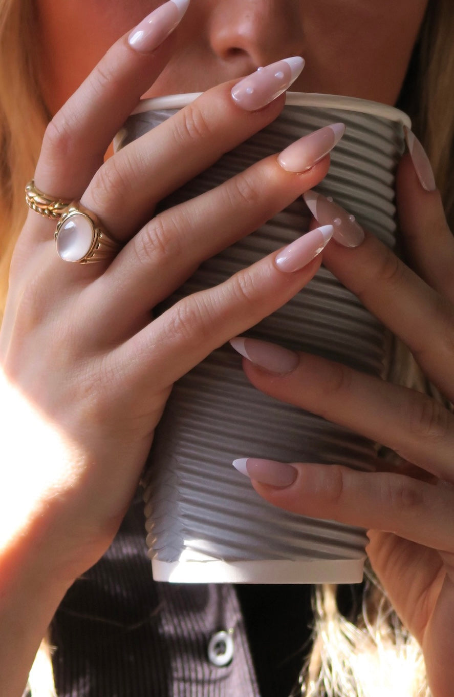 Close-up of hands wearing French tip press on nails with pearl detail holding a textured beige clutch with a blurred background