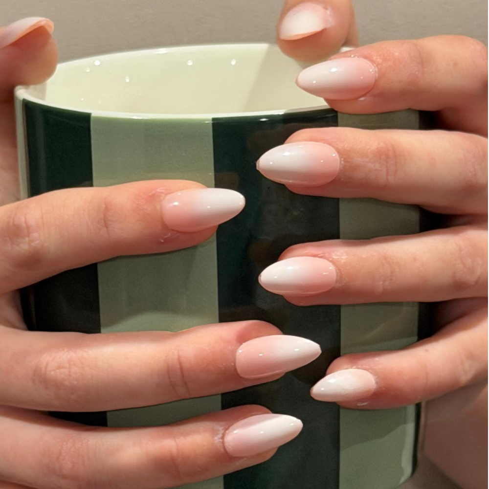 A close-up of hands holding a green striped ceramic mug, showcasing long, almond-shaped press-on nails from Sugar Tips with a glossy white-to-pale-pink ombre gradient.