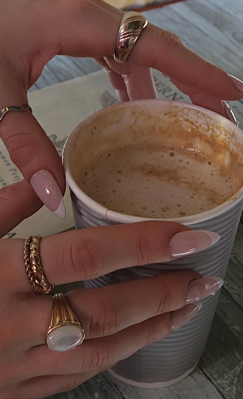 Person holding a paper cup with a partially consumed beverage, wearing gold rings and French tip press on nails with pearl detail