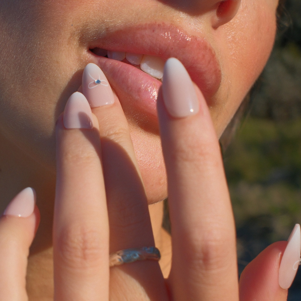 Close-up of a person's lips with a hand gently touching them, against a blurred natural background. Hands are wearing pink ombre press on anils with rhinestone detail