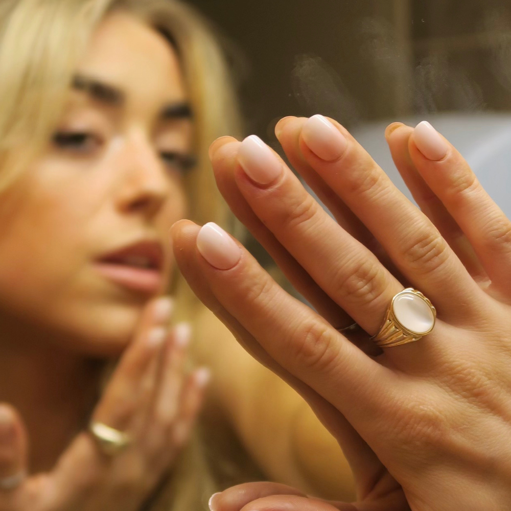 Woman wearing a gold ring with a large stone, looking at her reflection in a mirror, wearing short pink ombre press on nails