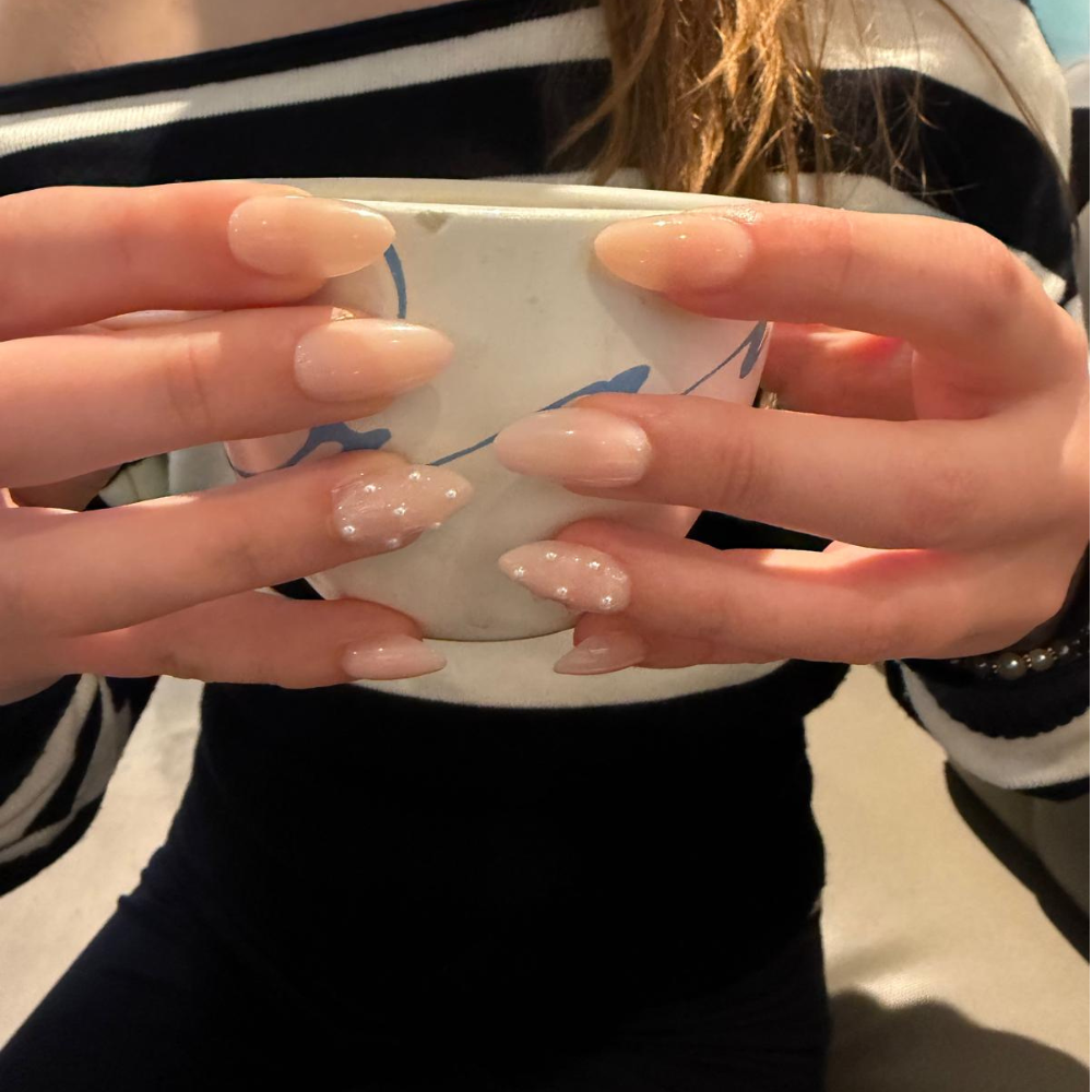 A close-up of a person's hands with long, glossy, almond-shaped press-on nails from Sugar Tips in a sheer nude-pink color with small white pearl accents on two of the nails, holding a white ceramic mug.