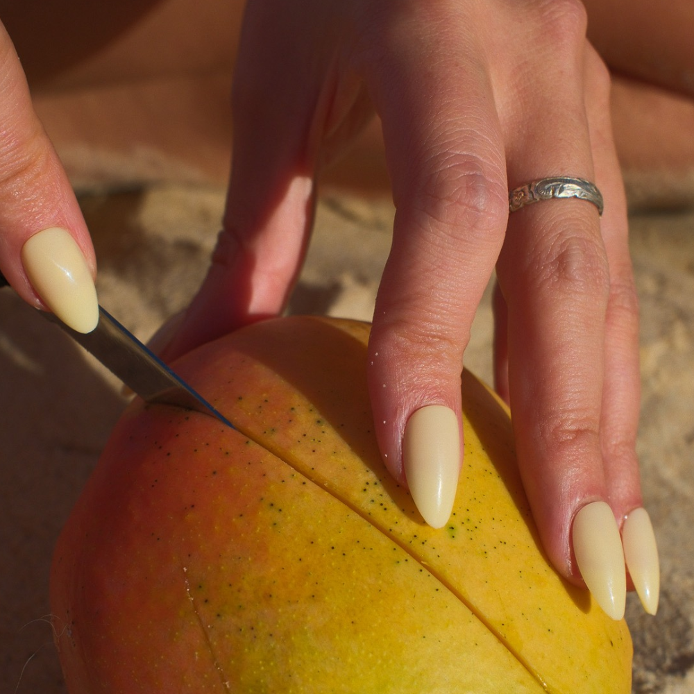 Butter Glow yellow almond press-on nails by Sugar Tips, slicing a mango with a knife on the sand
