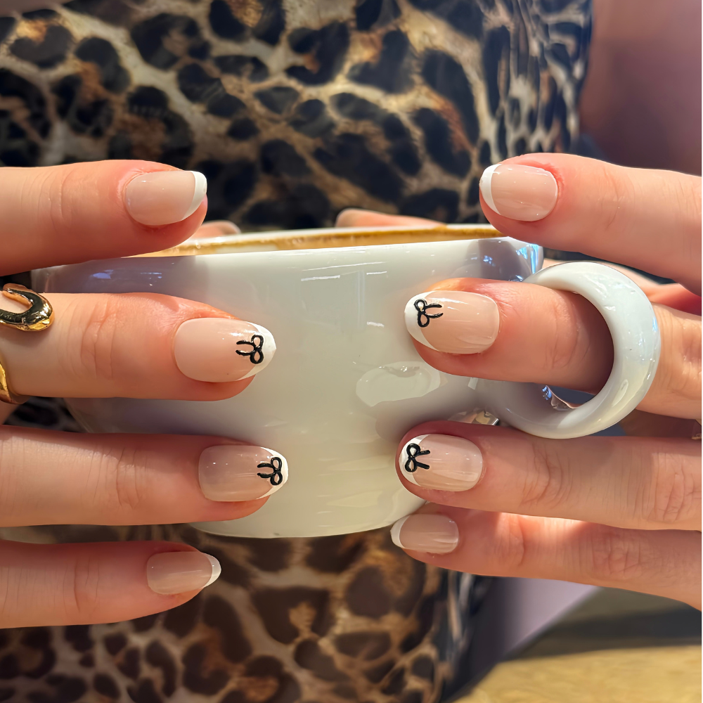 Close-up of hands holding a white ceramic mug, showcasing press on nails from Sugar Tips with a nude base, classic french tip, and small black bow details on the nails. The person is wearing a leopard print top and a gold ring on one finger.