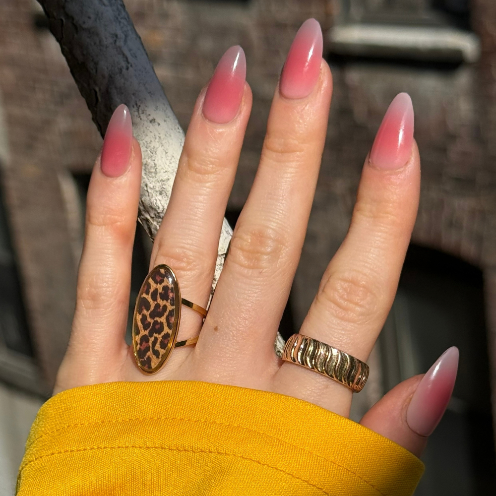 A close-up of a hand with pink ombre press on nails in long almond from Sugar Tips, wearing a leopard-print ring and a textured gold ring, with a yellow sleeve visible at the wrist.