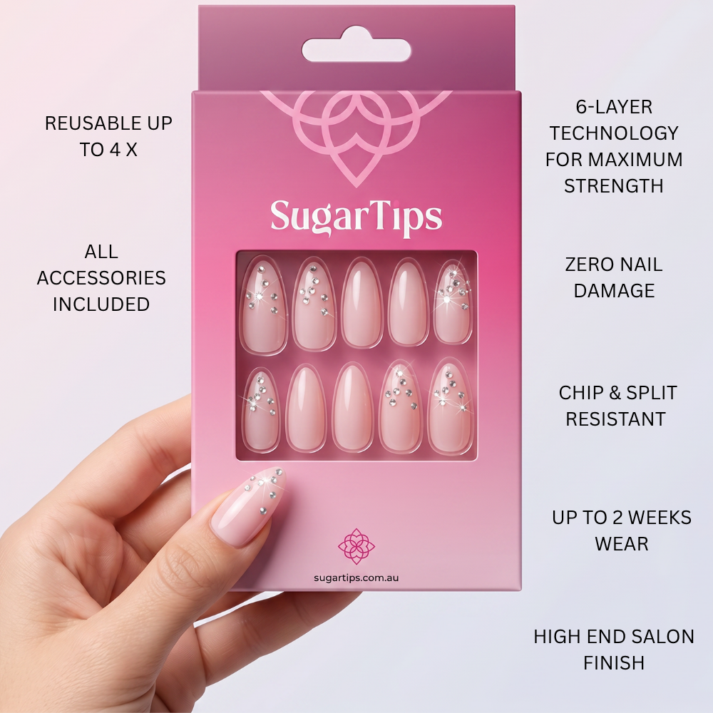 A hand holds a pink SugarTips press-on nail box against a pale pink background. The box shows ten almond-shaped nails with a glossy pale pink base and small silver rhinestone accents.